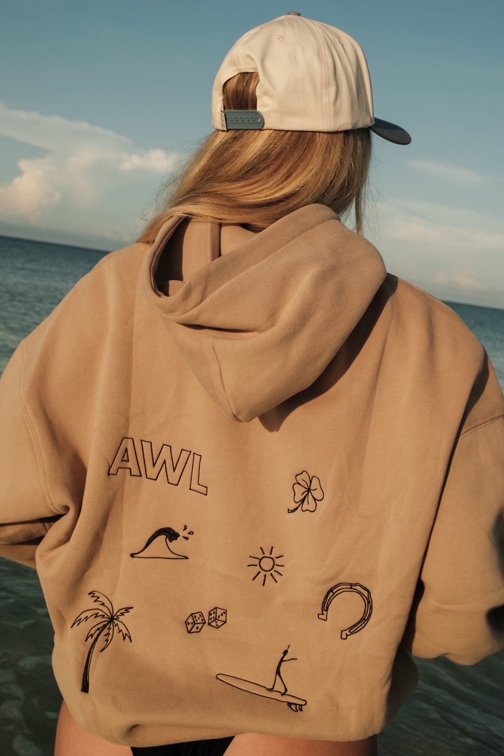 Girl wearing a beige hoodie with 'AWL' and various graphics, standing by the ocean.