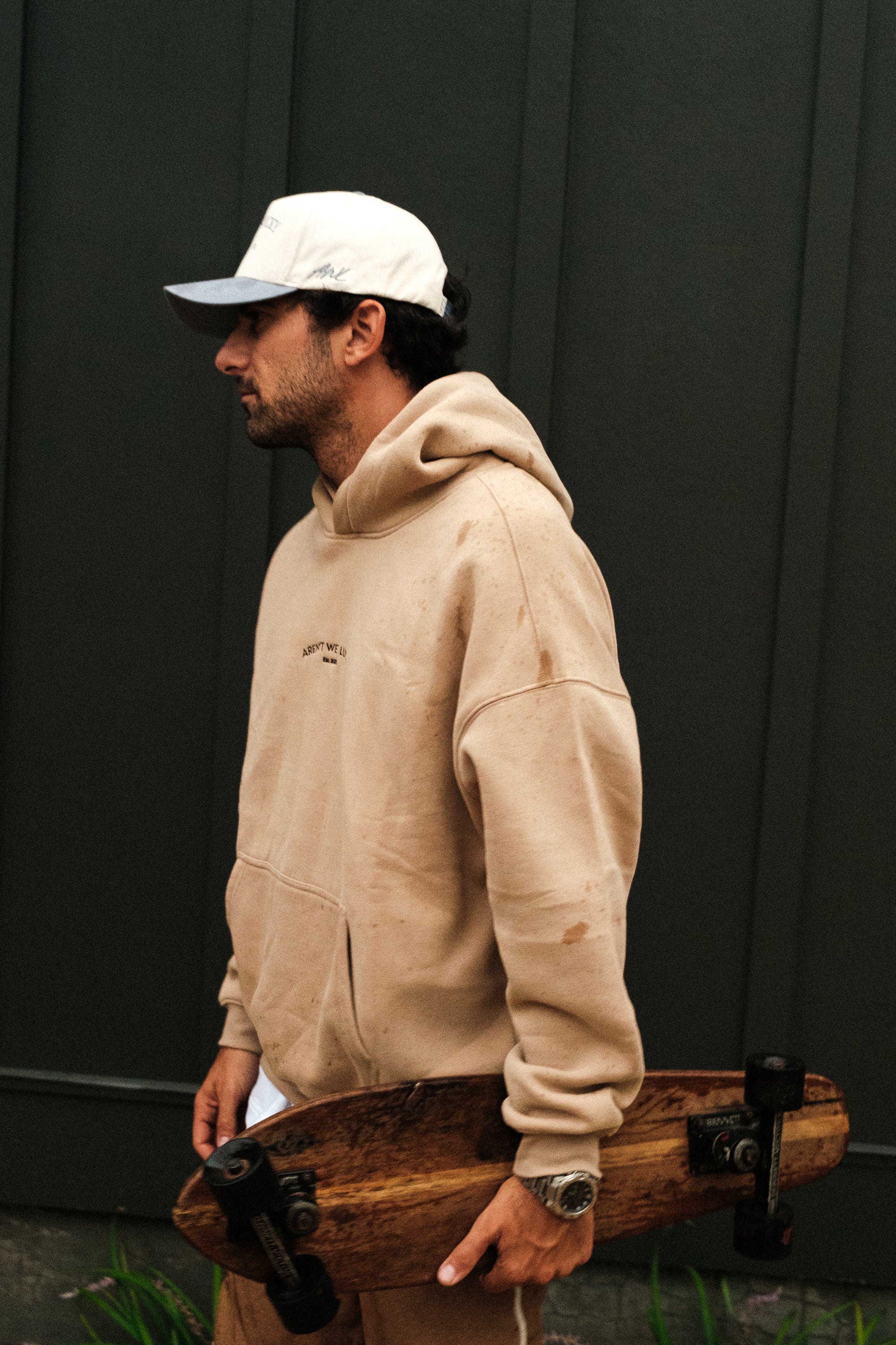 Man wearing a beige hoodie and white cap holding a wooden skateboard against a dark background