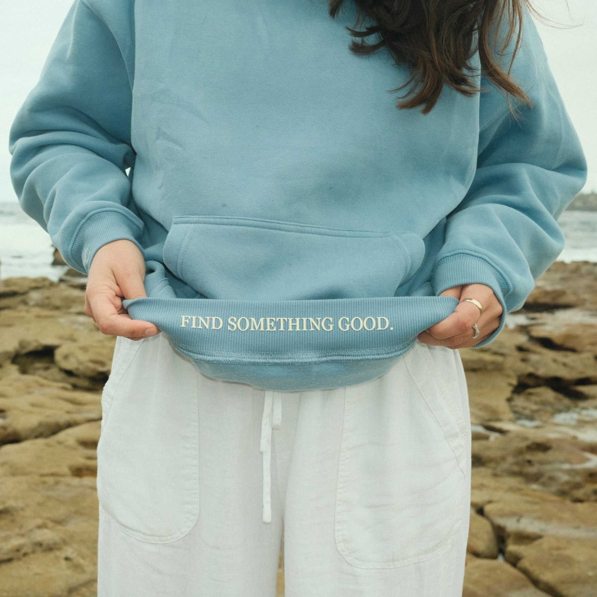Person wearing a blue hoodie with 'Find Something Good' text on a rocky beach.