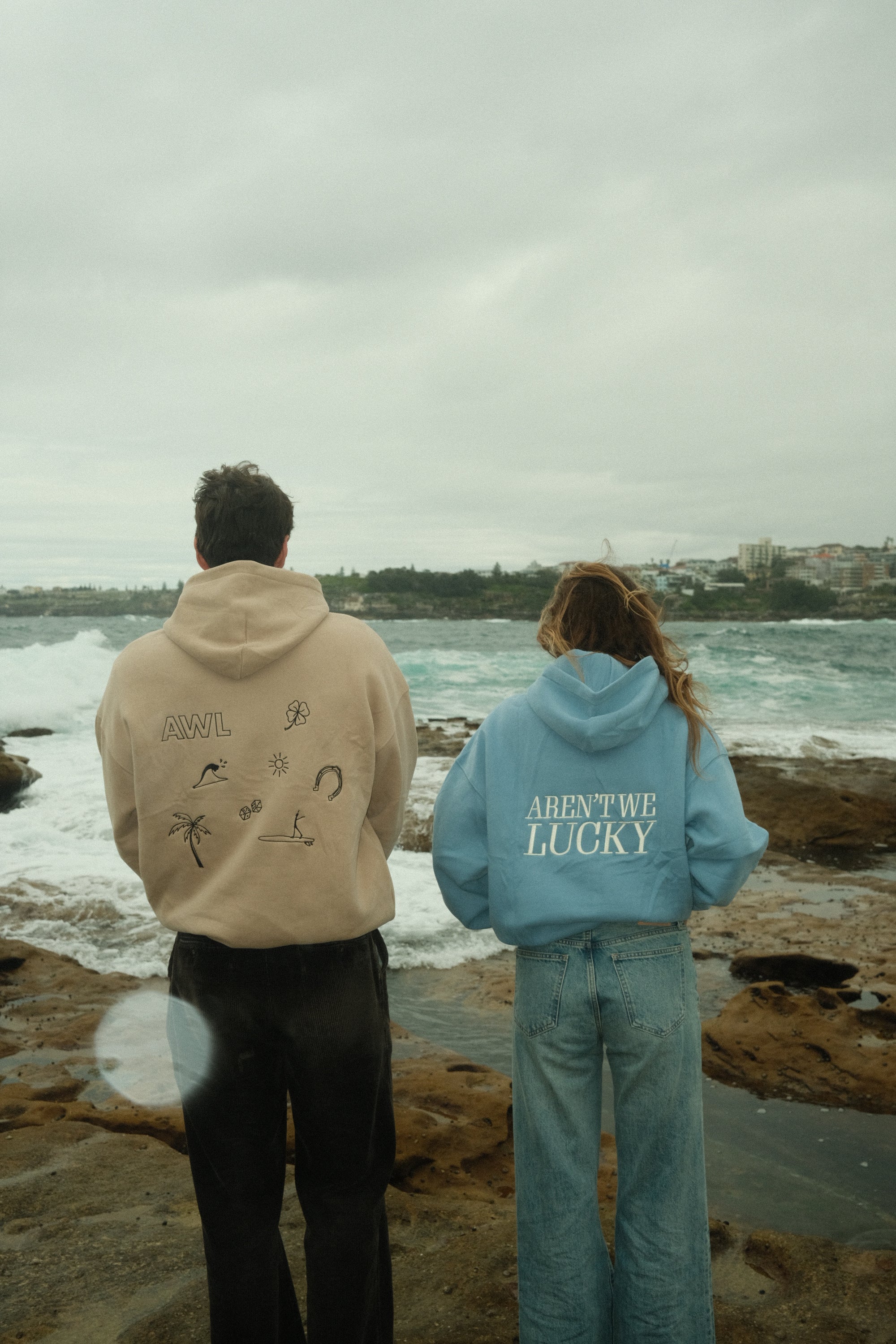 Two people wearing Aren't We Lucky sweatshirts with text standing on a rocky beach.