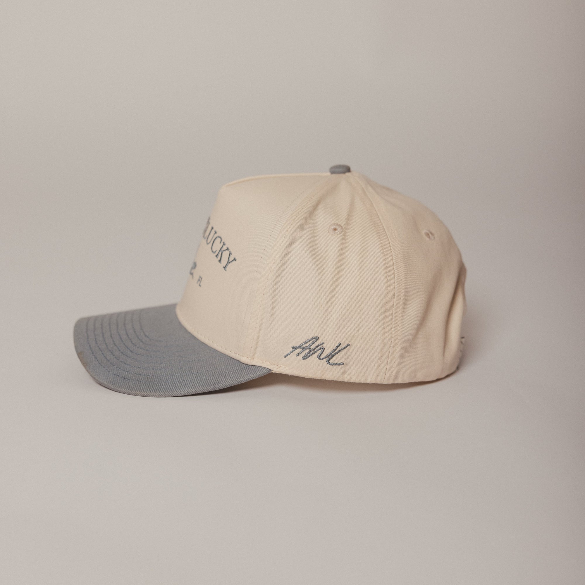 Beige and dusty blue Aren't We Lucky cap with text and logo on a beige background AWL side