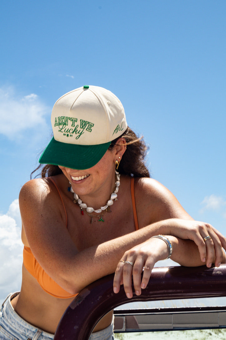 Woman wearing a two tone Aren't We Lucky cap with text, sitting by a car against a blue sky.
