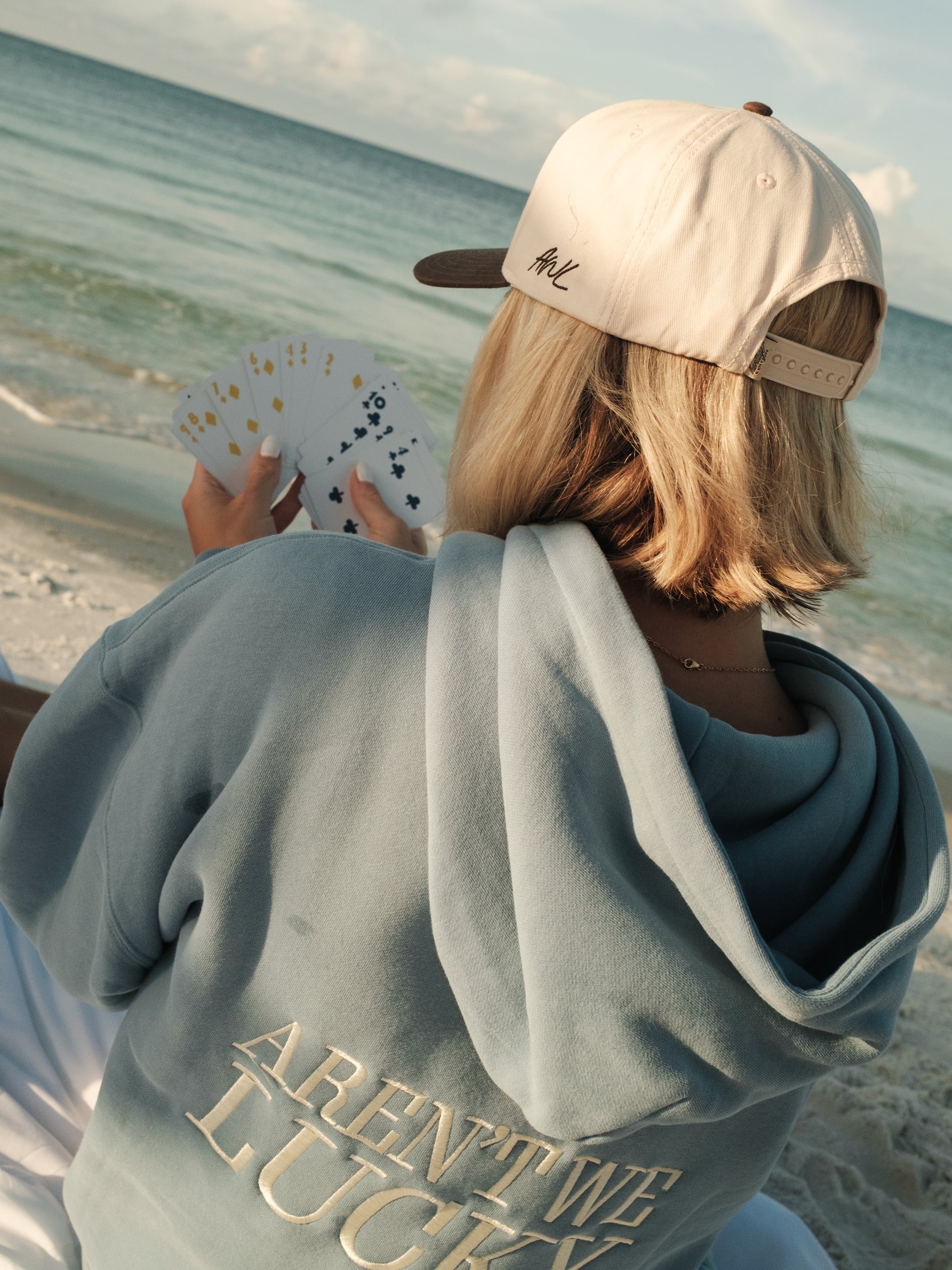Person sitting on a beach holding playing cards, wearing a cap and a hoodie with text.
