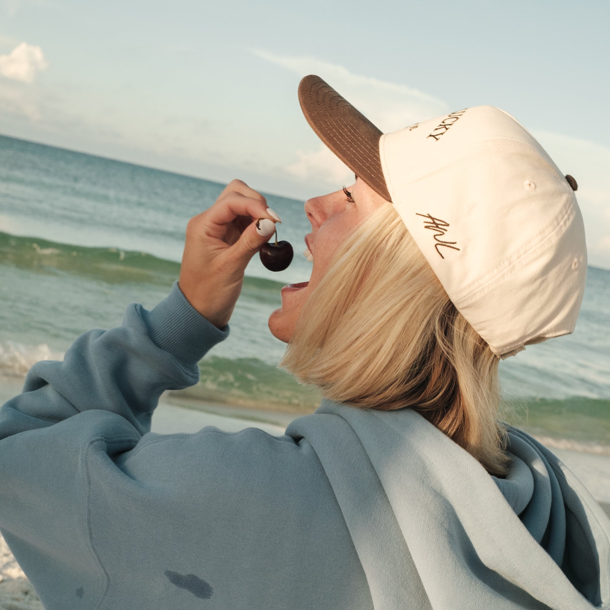 Lifestyle photo of person wearing an Aren't We Lucky cap and hoodie by the beach