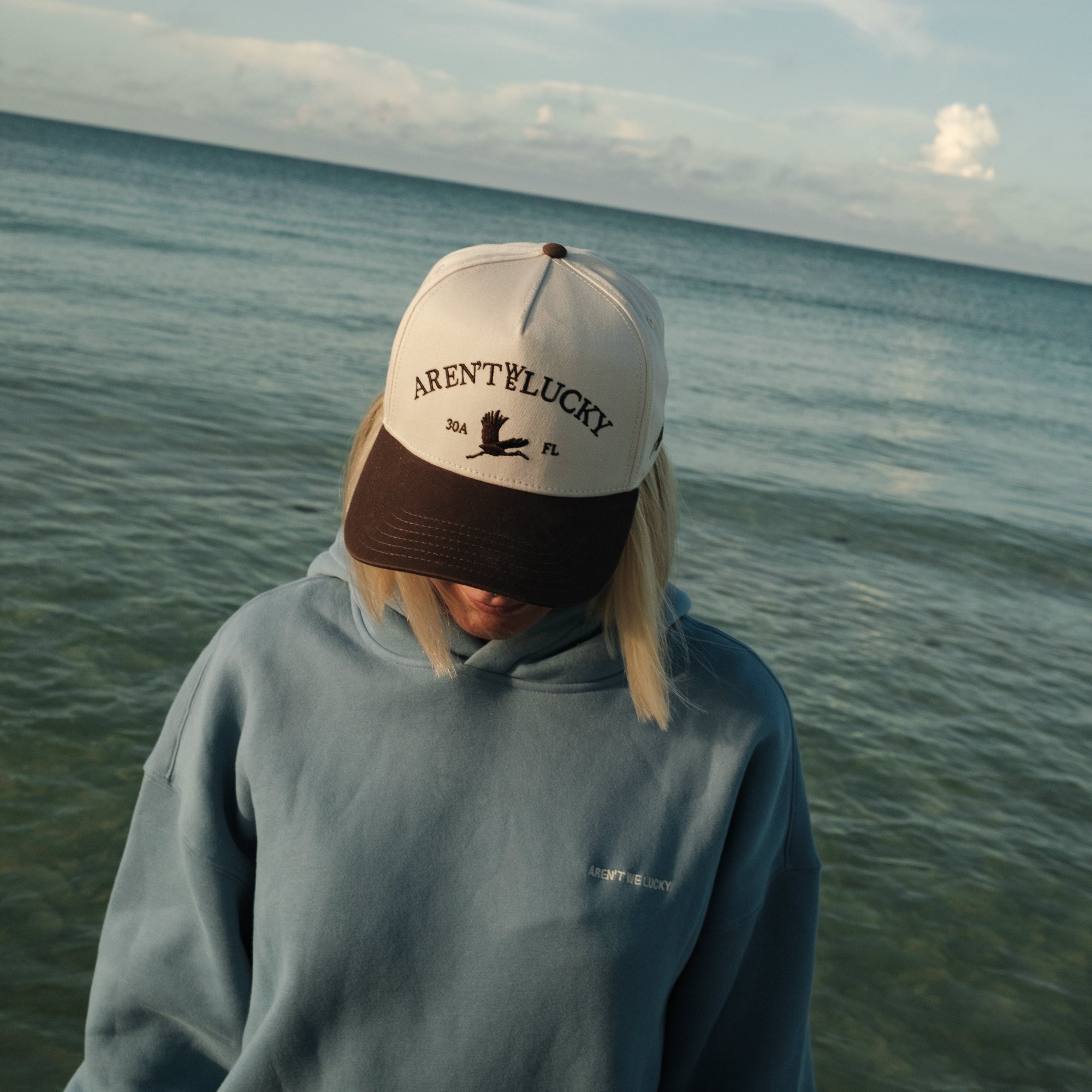 Person wearing an Aren't We Lucky cap with text standing by the ocean