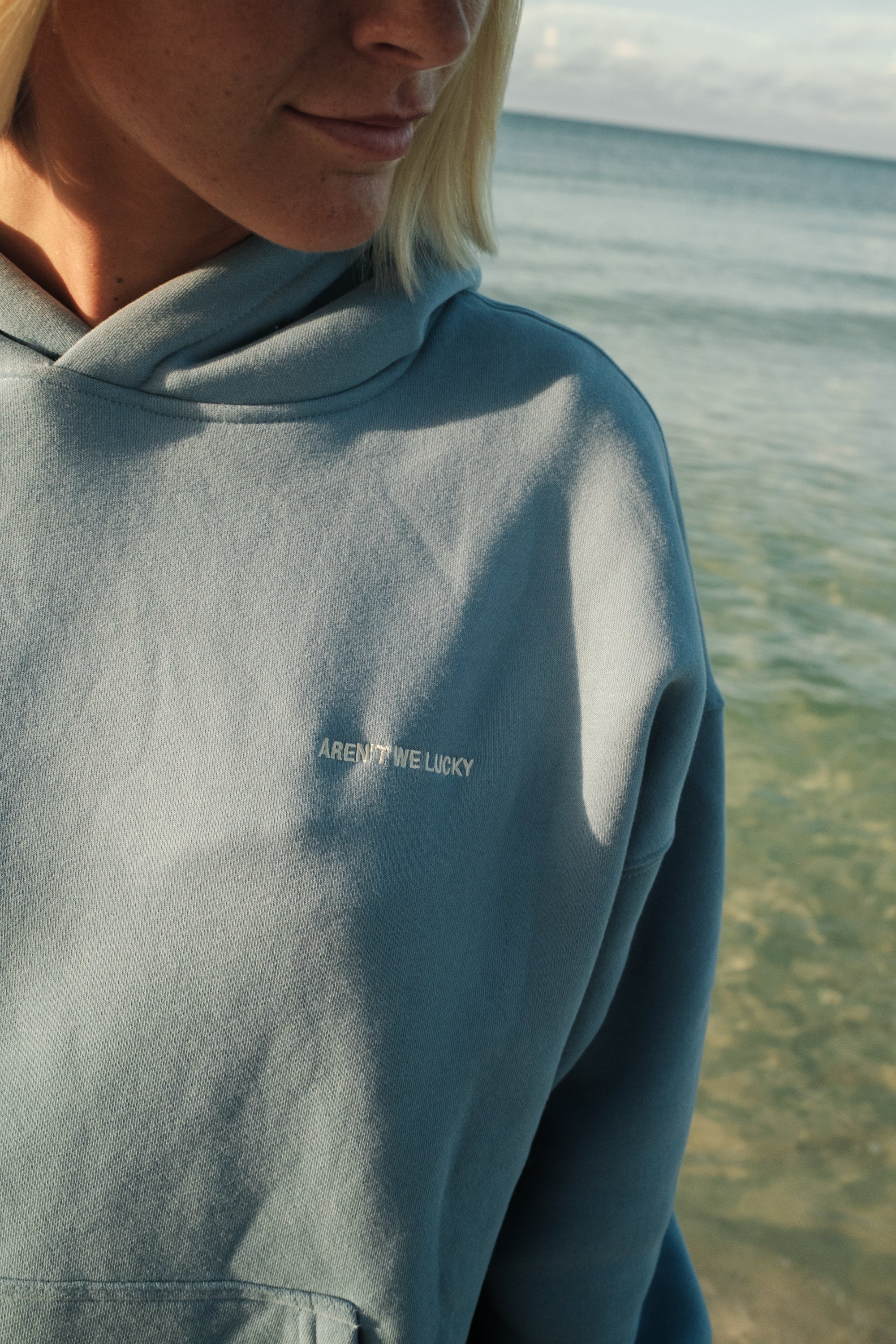 Person wearing a hoodie with 'ARE WE THE LUCKY' text by the water