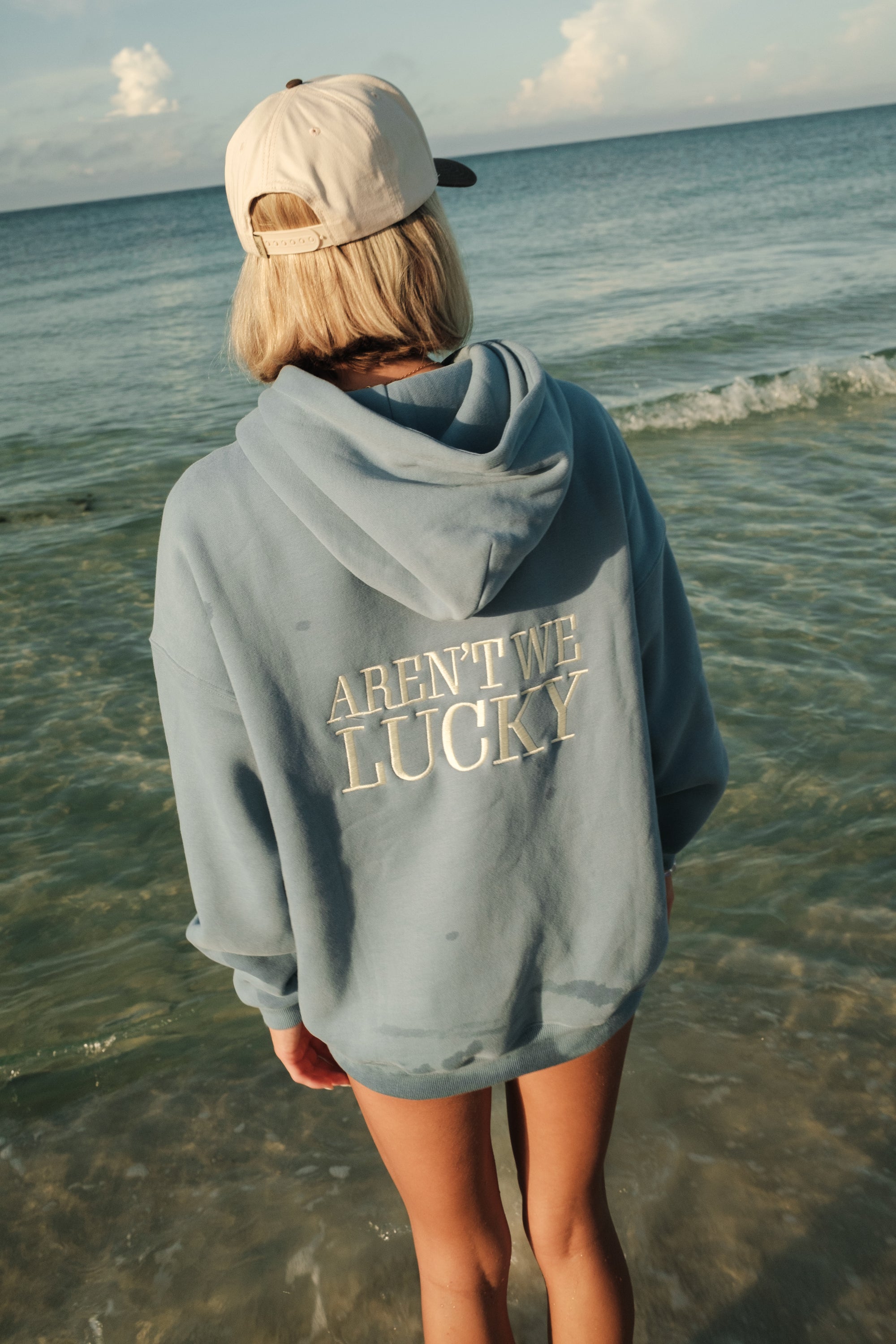 Person wearing a hoodie with 'Aren't We Lucky' text on a beach