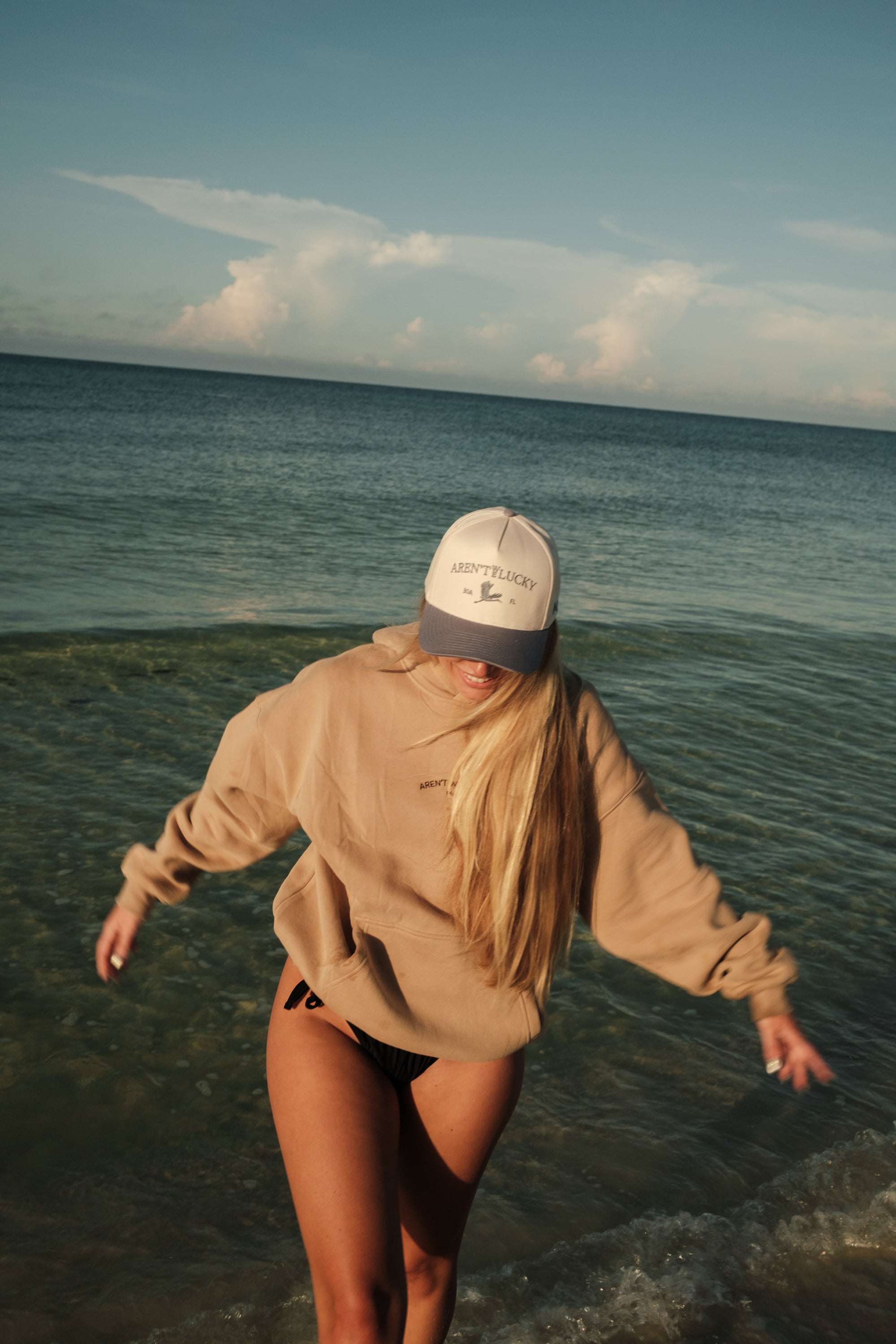 Person wearing a beige hoodie and cap walking along a beach with ocean waves.