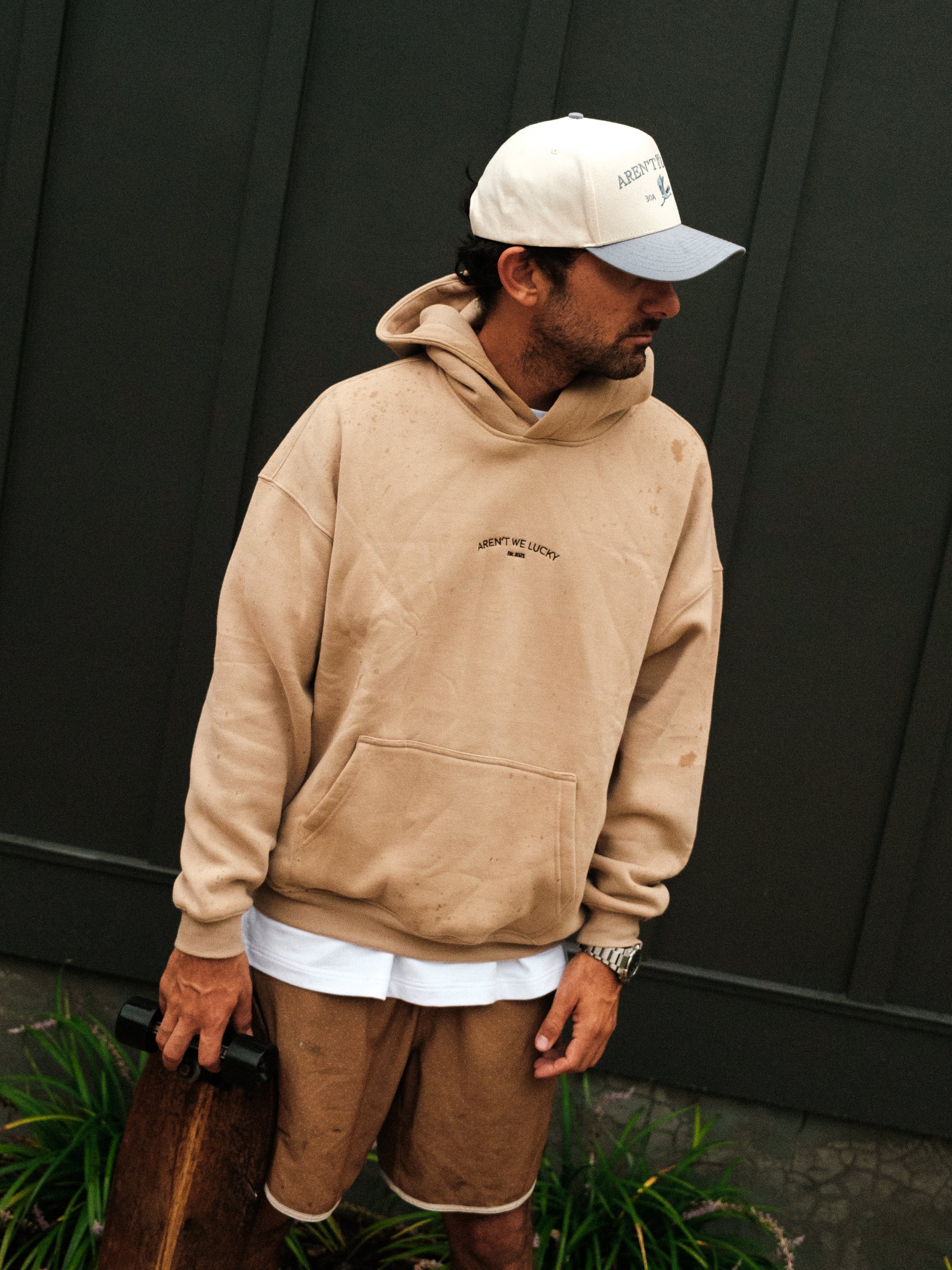 Man wearing a beige hoodie and shorts with a cap, standing against a dark wall.