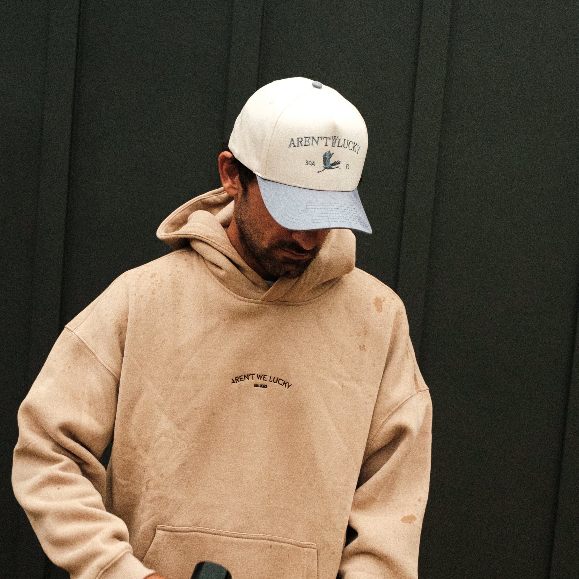 Person wearing a beige hoodie and cap holding a skateboard against a dark background