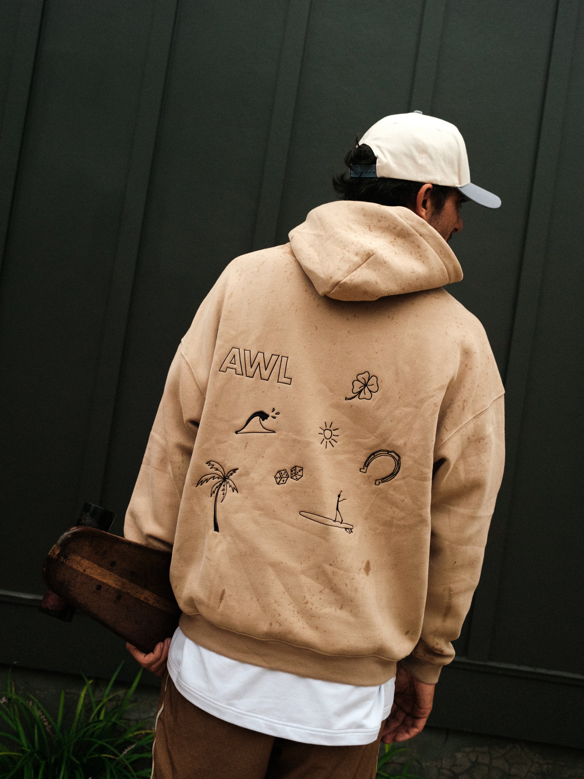 Person wearing a beige hoodie with various designs and 'AWL' branding, standing against a dark wall.