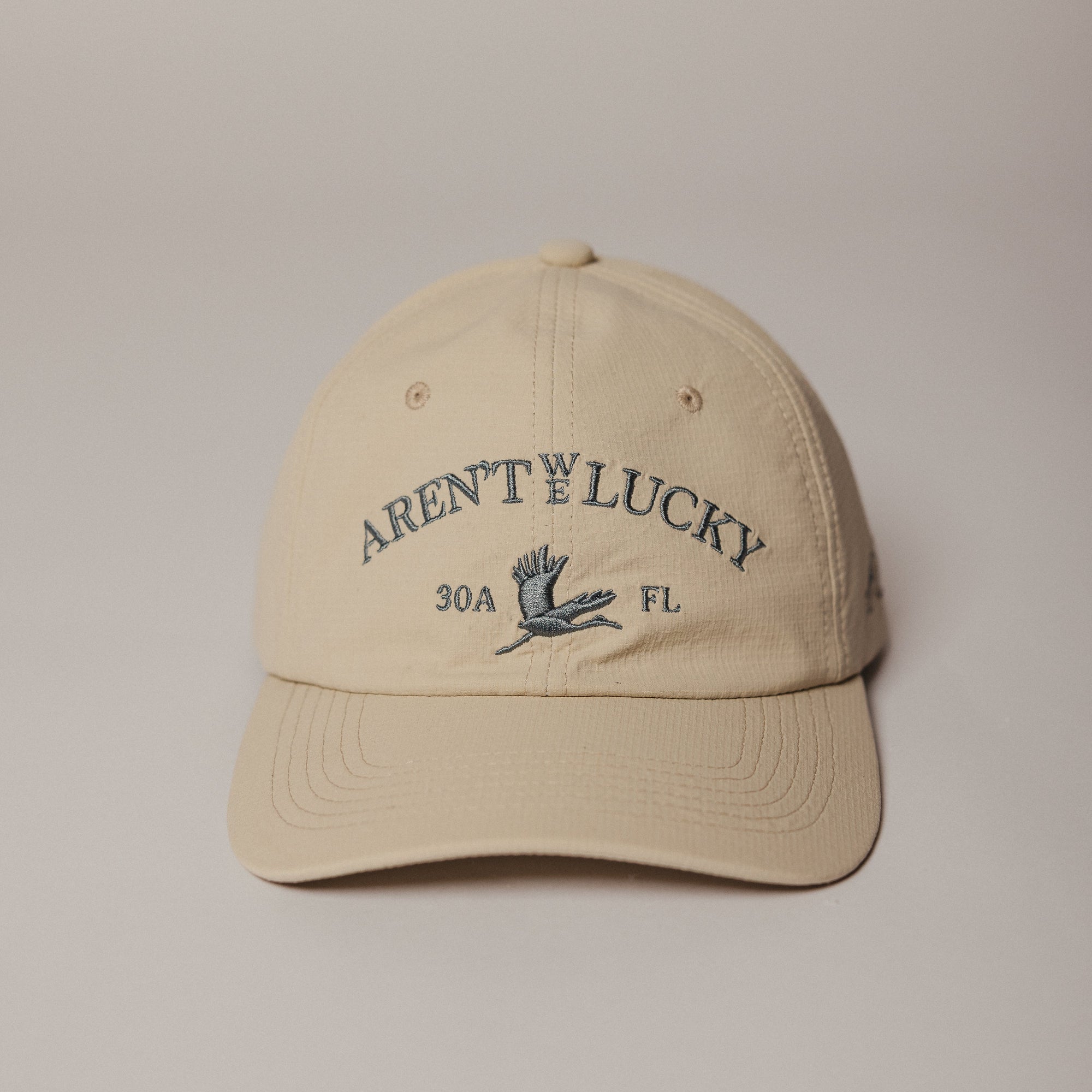Beige Aren't We Lucky 30a Crane Dry Baseball cap with text and logo on a beige background