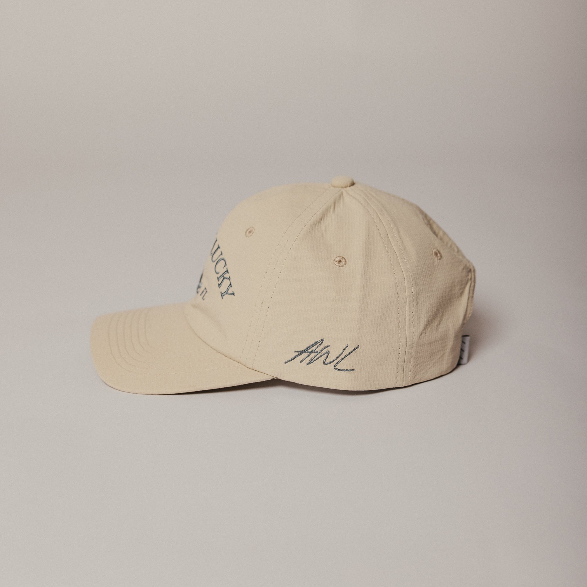 Beige Aren't We Lucky 30a Crane Dry Baseball cap with text and logo on a beige background. AWL on side