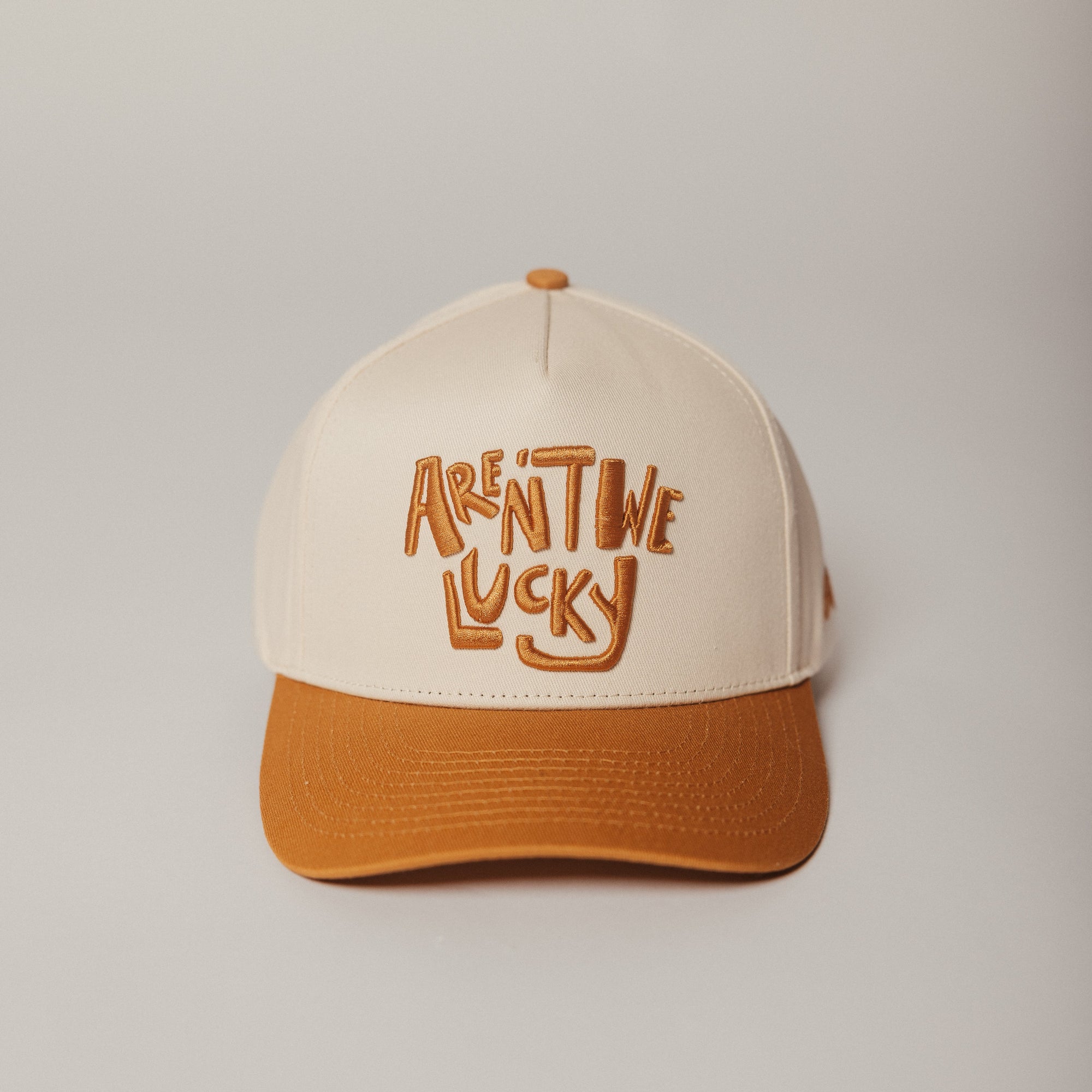 Beige and orange Aren't We Lucky trucker cap with text on a light gray background