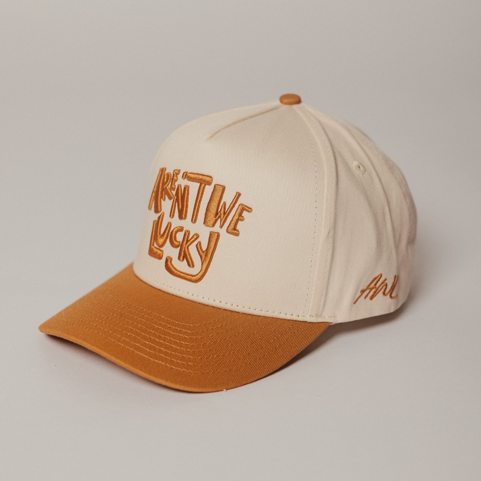 Beige and orange Aren't We Lucky trucker cap with text on a light gray background